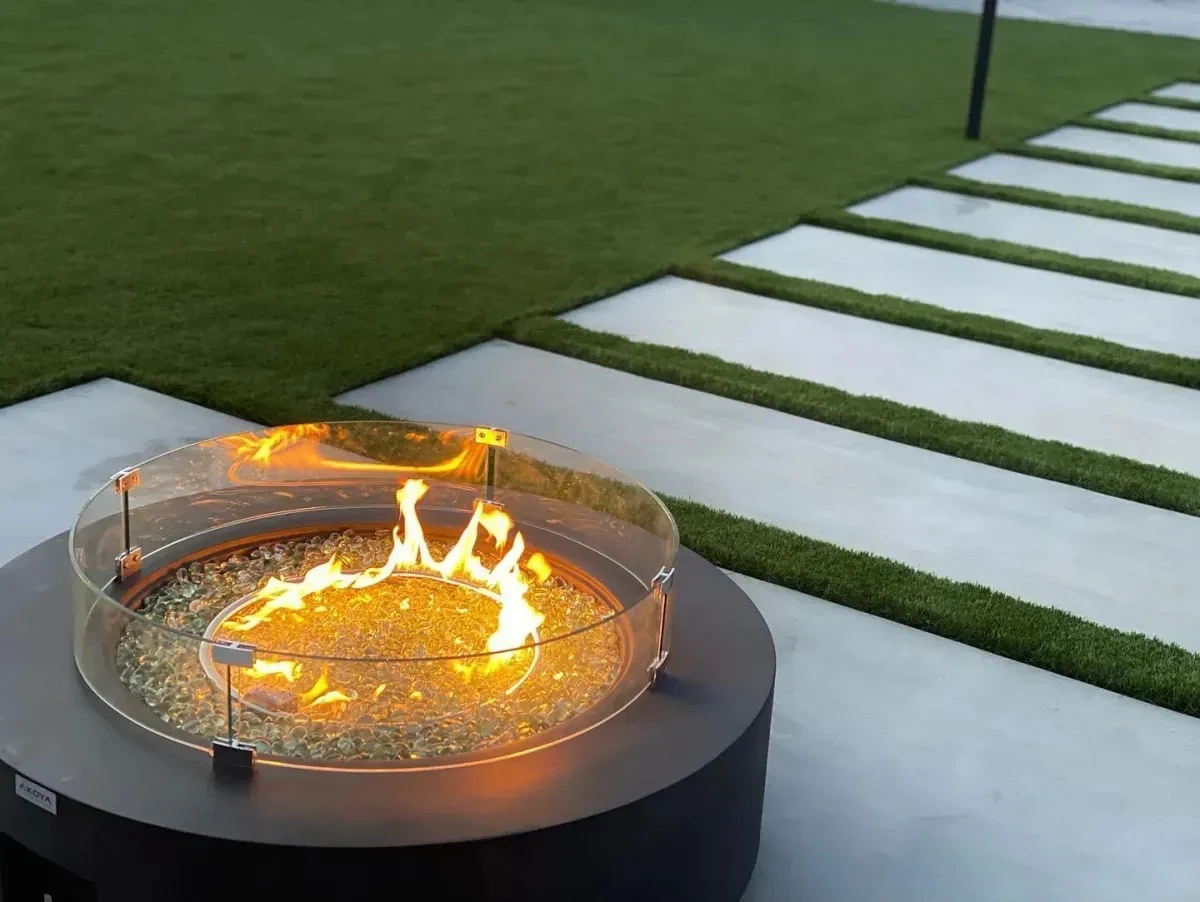 Round gas fire bowl burning at dusk on artificial turf beside concrete stepping panels and a path light in Orange County, California