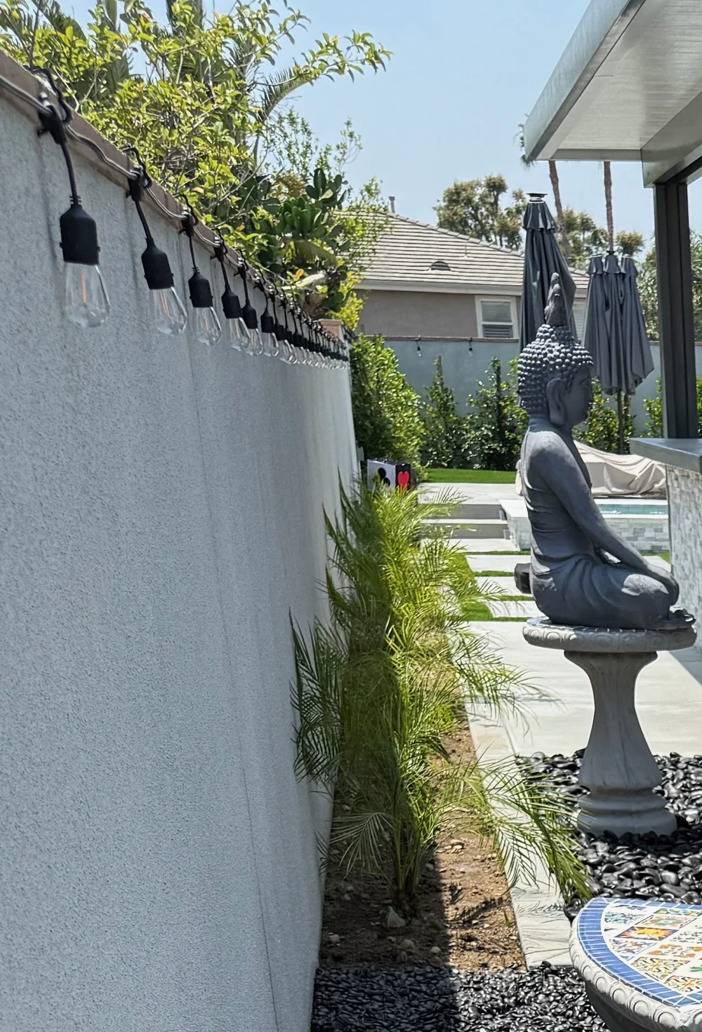 Concrete stepping panels with turf joints crossing a black pebble side yard with new palm planting and string lights in Orange County, California